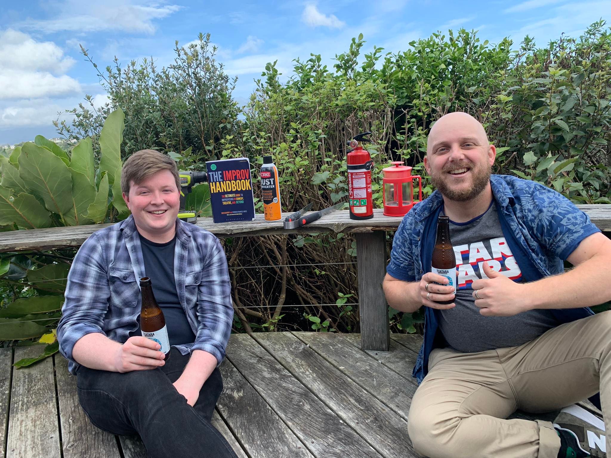 Austin and Dylan, two young white men, sit on a deck with beers and camping equipment. THere is a copy of The Improv Handbook on the deck with them.