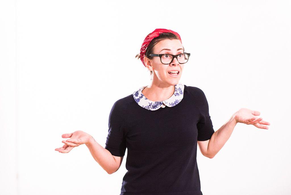 A photo of Andel. She is a white woman wearing a red hair scarf and black dress, looking off camera with her hands in a shrug